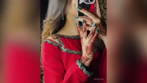 Woman in elegant red traditional outfit with intricate henna patterns and teal nail polish taking a selfie.