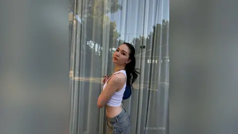 Stylish young woman in a white tank top and denim jeans posing by a window with sheer curtains in soft daylight.