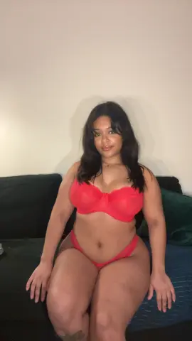 Hazelvalentine wearing a red bra and matching red thong, sitting on a dark couch against a plain wall.