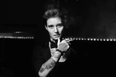 Moody black and white portrait of a tattooed man in a black shirt holding a cigarette in a dark setting.