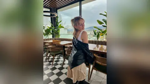 Stylish woman in a black leather jacket and white tank top posing in a modern rooftop restaurant.