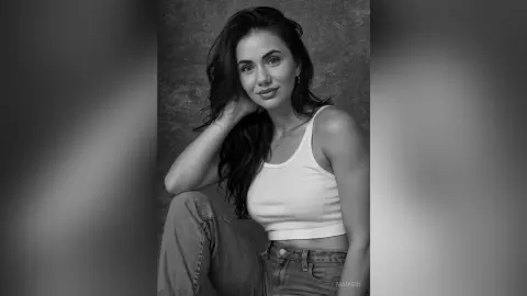 Smiling young woman in a white tank top and jeans posing against a textured studio background in black and white.