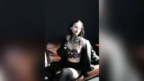 KailaTaylor poses sitting on a leather sofa wearing a black lace bra, black skirt, and black jacket with her dark hair down.