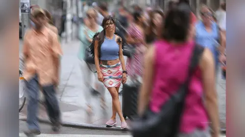Stylish woman in a blue tank top and patterned skirt walking through a busy, sunlit city street.