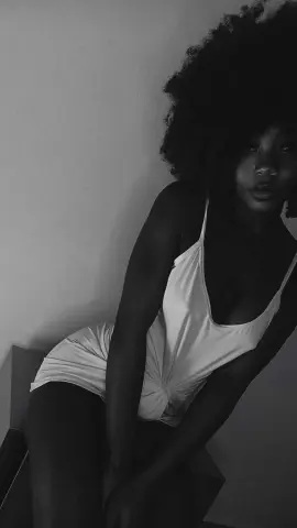 Black woman in a white bodysuit posing against a minimalist wall in a moody black and white portrait.