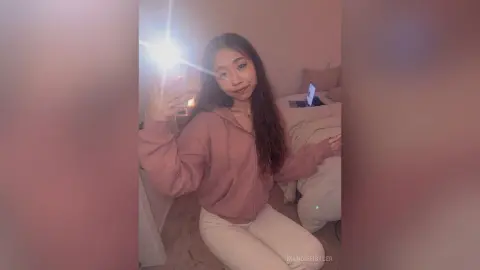 Young woman in a pink hoodie relaxing in a cozy bedroom with soft, warm lighting.