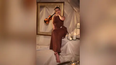 Elegant woman in a brown satin dress posing with a violin in a moody, candlelit room.