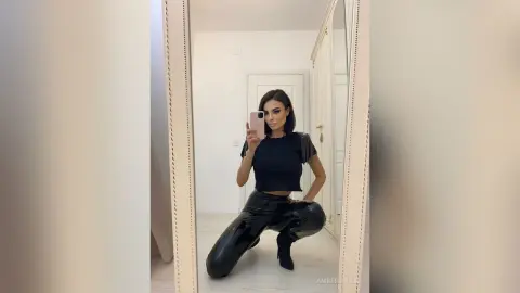 Stylish woman in a black crop top and shiny patent leather pants posing for a mirror selfie in a bright room.