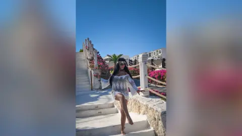 MissyJolie stands on white stone stairs wearing a white lace cover-up and sunglasses against a clear blue sky.