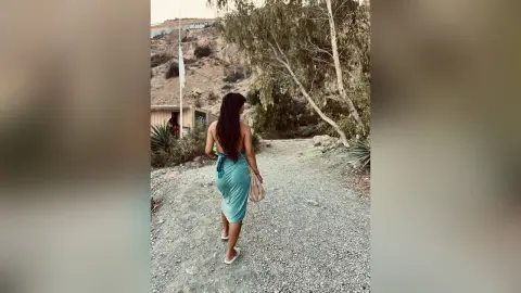 Woman in a teal summer dress walking down a dusty mountain path toward a scenic landscape.