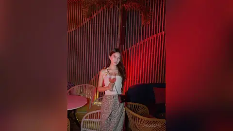 CheCascano poses in a white tank top and patterned pants while holding a drink at an outdoor lounge.