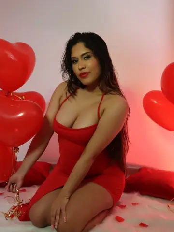 Young woman in a red mini dress sitting among red heart balloons in a romantic indoor setting.