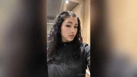 Young woman with long curly hair wearing a black zip-up jacket in a modern indoor setting.