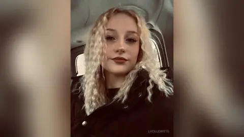 Blonde woman with wavy hair wearing a black jacket sitting in a car with a moody aesthetic.