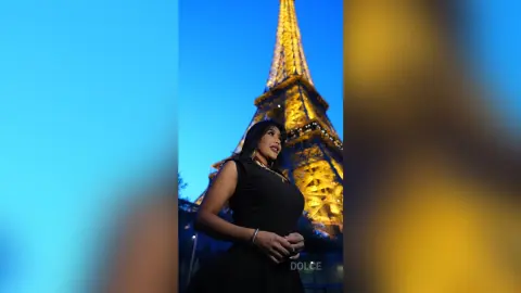 Dolce poses in a black sleeveless dress against the lit Eiffel Tower at night.