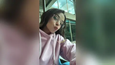 MielVerane pouts her lips while wearing glasses and a light pink hoodie in an indoor setting.