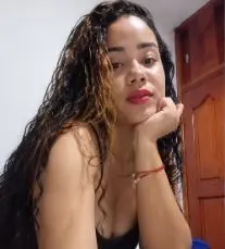 Luciana_Bloom poses with her hand under her chin, showing her long wavy hair and red lipstick while wearing a black top.