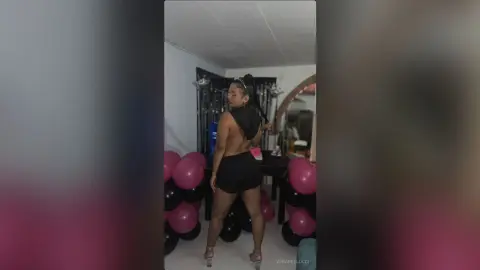 Confident woman in a black backless top and shorts posing in a room with pink balloons.