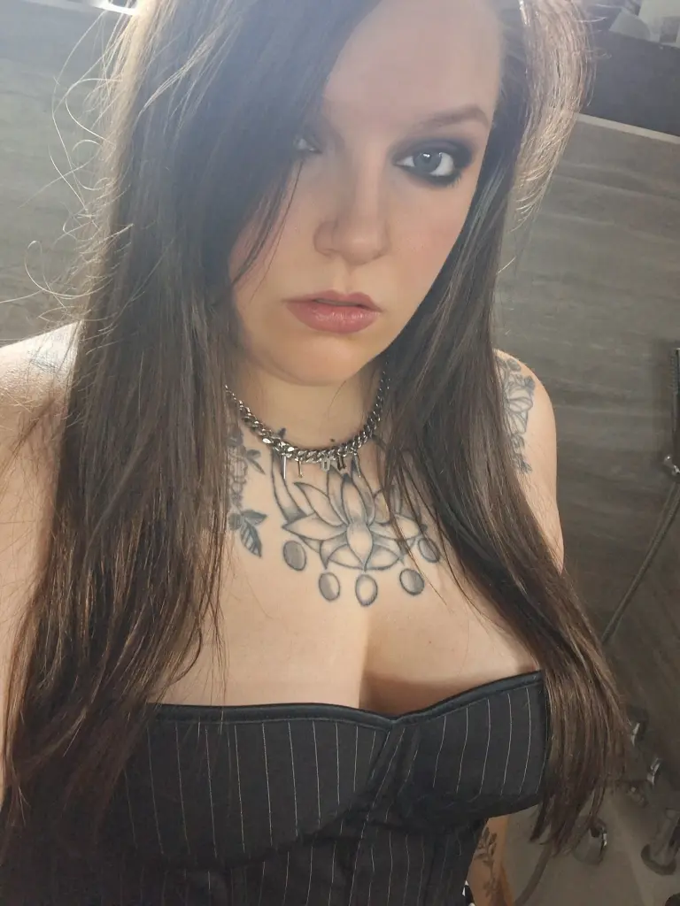 Gothiccbxby poses in a black pinstripe corset, showing off her cleavage and long dark hair against a neutral background.