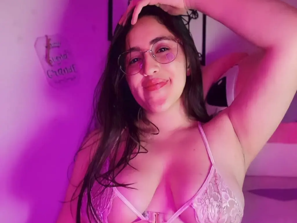 RosaWeyr smiles while wearing glasses and a white lace bra, posing with one hand behind her head in pink lighting.