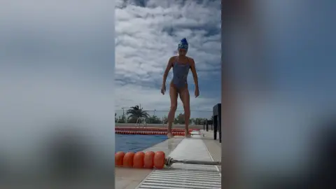 Young woman in a patterned swimsuit standing on a pool diving board under a cloudy sky.