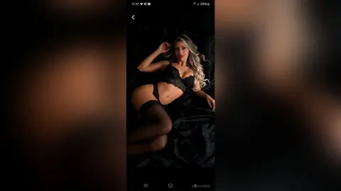 Blonde woman in black lace lingerie lounging on dark satin sheets in a moody, intimate bedroom setting.