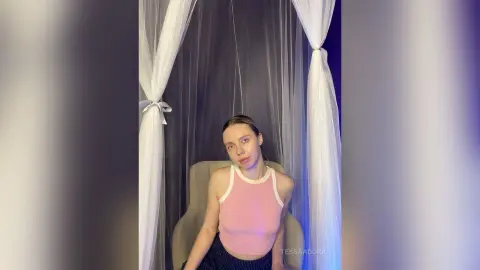 Young woman in a pink halter top sitting in a dreamy white curtain tent setting with soft lighting.