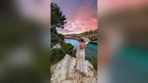 Woman in a white maxi dress standing on a rocky cliff overlooking a turquoise ocean at sunset.