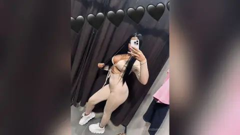 Young woman in a beige cutout bodysuit and white sneakers posing for a mirror selfie in a fitting room.