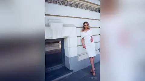Elegant woman in a white cocktail dress posing against a classic stone building in soft daylight.