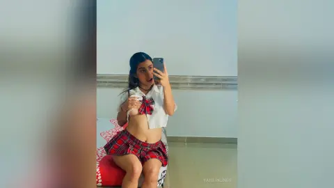 Young woman in a red plaid schoolgirl outfit taking a mirror selfie in a bright room.
