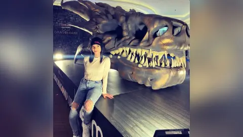 Woman in white long-sleeve shirt and jeans sitting next to a massive dragon skull sculpture in a dark museum.