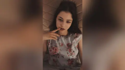 Young woman in a floral print blouse lying on a bed with a dreamy, moody expression.