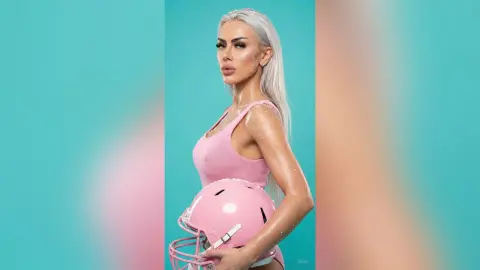 Blonde woman in a pink tank top holding a pink football helmet against a turquoise background.