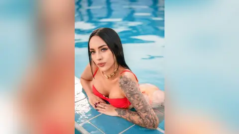 Tattooed woman in a red bikini relaxing in a bright blue swimming pool on a sunny summer day.