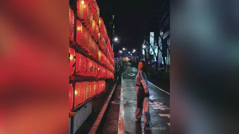 AnaMathers stands on a dark street next to a wall of red lanterns, wearing a blue and white striped long-sleeve top and dark pants.