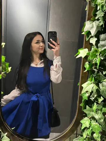 Woman in a blue dress and white blouse taking a mirror selfie near green ivy leaves.
