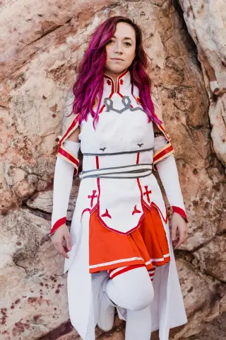 Woman in white and red fantasy cosplay outfit standing against a rugged rock wall in a natural outdoor setting.
