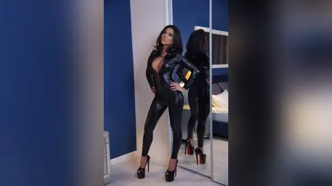 KinsleyCatalina poses in a tight black leather jumpsuit and high heels against a blue wall.
