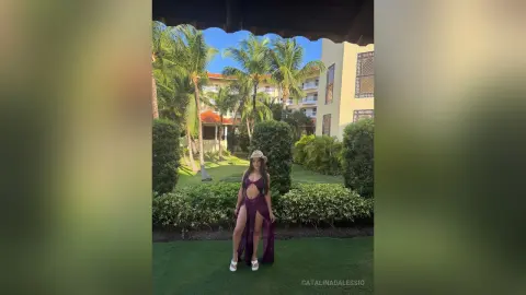 Woman in a purple cutout maxi dress and sun hat relaxing in a lush tropical garden resort.