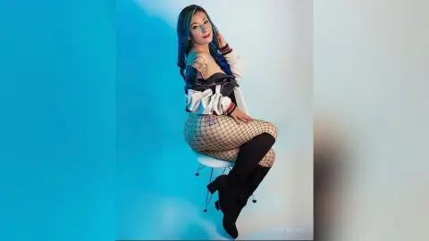 Woman with blue hair in a white top and fishnet tights posing against a blue studio background.