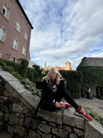 NikaZen sits on a stone wall outdoors wearing a black hoodie and black leggings with red sneakers.