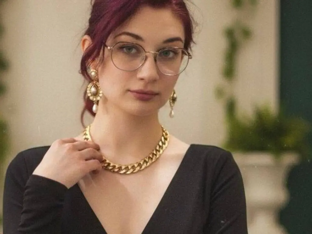 Elegant woman with red hair and gold jewelry wearing a black V-neck top in a soft indoor setting.