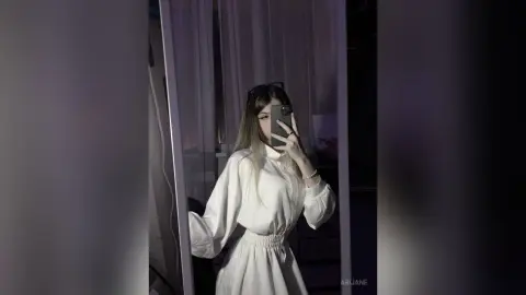 Young woman in a white long-sleeve blouse posing for a moody mirror selfie in a dimly lit room.