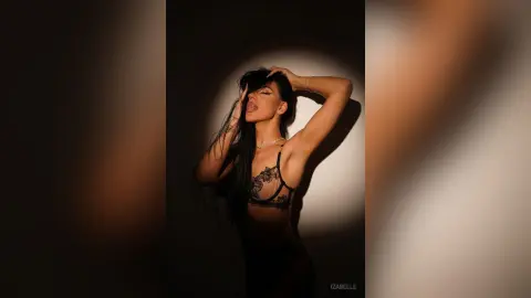 Iza — Young woman in black lace lingerie posing with long dark hair against a moody, shadowed studio background.