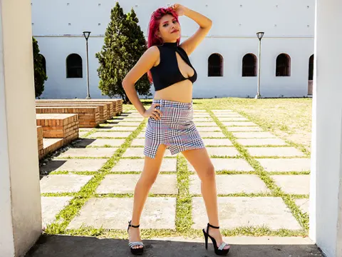 Red-haired woman in a black halter top and plaid mini skirt posing in a sunny outdoor courtyard.