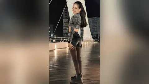 Smiling young woman in a patterned crop top and black shorts posing on a wooden boardwalk at night.