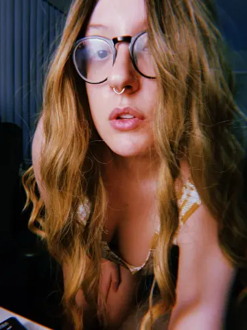 Young woman with long blonde hair and round glasses wearing a white patterned top in a soft indoor setting.