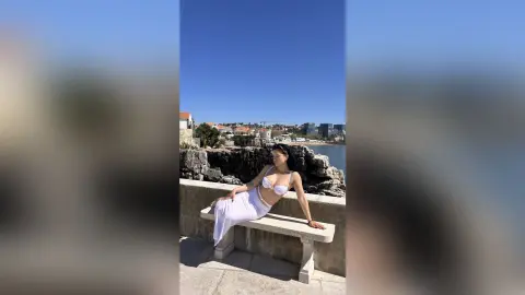 Young woman in a white bikini top and long skirt sitting on a stone bench by the rocky seaside under a blue sky.