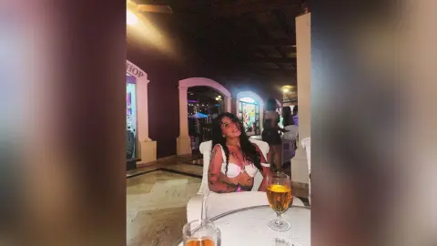 RossiNoleen smiles while wearing white lace lingerie and sitting behind a glass of amber liquid on a white table.